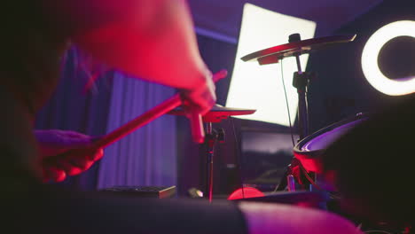 close up of lady playing electronic drum set, striking cymbal while pressing pedal, under moody red and purple lighting with drum pads, monitor screen, and ring light visible in indoor studio setup