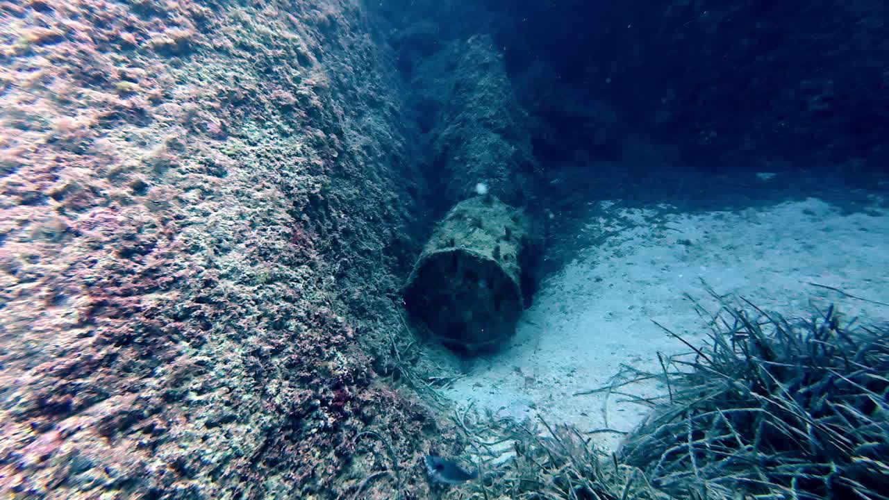 Ripresa subacquea cinematografica di un enorme barile di ferro arrugginito incrostato di vegetazione marina adagiato sul fondale sabbioso di una profonda fossa mediterranea blu vicino a Malta durante una limpida mattina d'estate