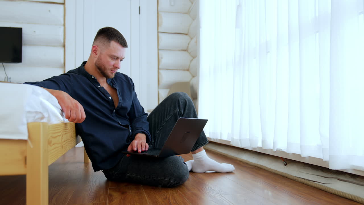 Man in dark blue shirt sits on the floor leaning on the bed. Bearded male holds a laptop on his knee.