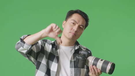 화가 난 아시아 사진작가가가 카메라에서 사진을 찍고, 그 다음에는 스튜디오에서 녹색 스크린 배경에 서서 엄지손가락을 내려놓는 제스처를 보여준다.