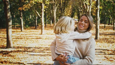 una joven madre rubia está sonriendo, sosteniendo a su hija en sus brazos y dando vueltas con ella durante una caminata en el parque de otoño