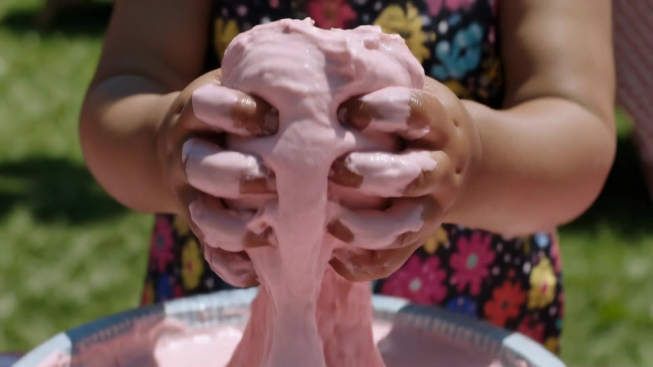 Bambina in vestito floreale che si allunga e preme dello slime da una ciotola a strisce su un tavolo a scacchi, giocando.