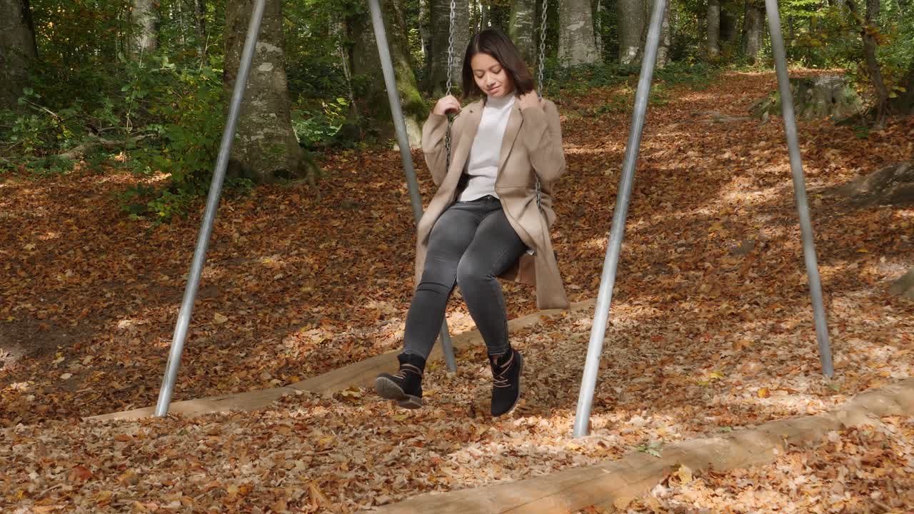 mujer bonita en columpio en el bosque durante el día soleado en otoño con hojas de colores que caen - tiro medio estático
