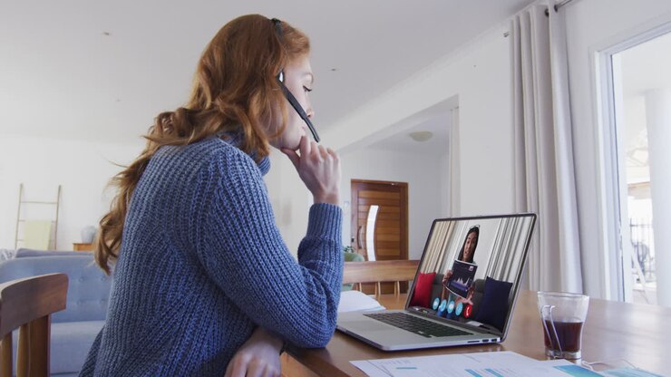 Donna caucasica con auricolare telefonico in videochiamata con una collega donna su laptop a casa.