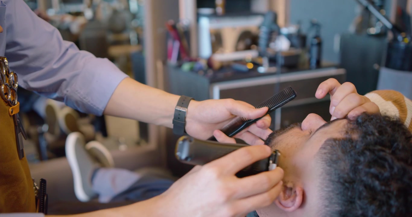Afroamerikansk barber trimmer skæg med trimmer, mens mandlig kunde læner sig tilbage i butikken for en skarp kant.