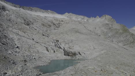 Drone-climbs-over-unique-glacial-lake-with-a-river-flowing-on-granite-stones,-shaped-by-the-Neves-Glacier-visible-in-the-background