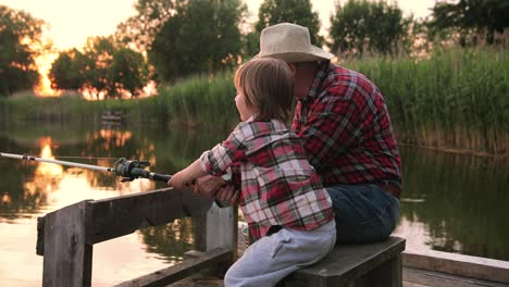 Rear-view-of-a-grandfather-and-his-grandson-catching-fish-sitting-on-the-lake-pier-on-a-summer-day-at-sunset