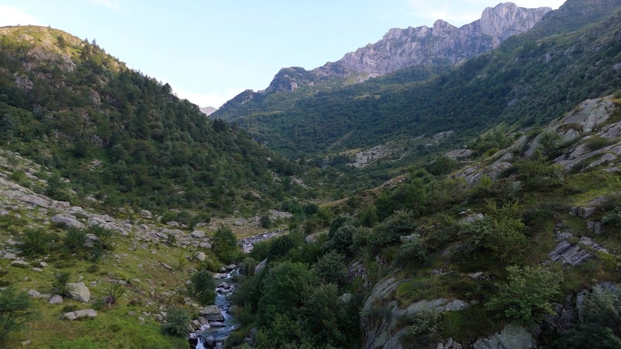 Un piccolo ruscello serpeggia attraverso una valle aspra e alberata sotto alte vette, mentre la telecamera aerea scivola lentamente in avanti