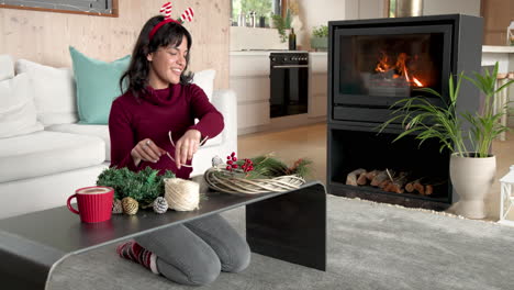 woman crafting christmas wreath at home, smiling by cozy fireplace