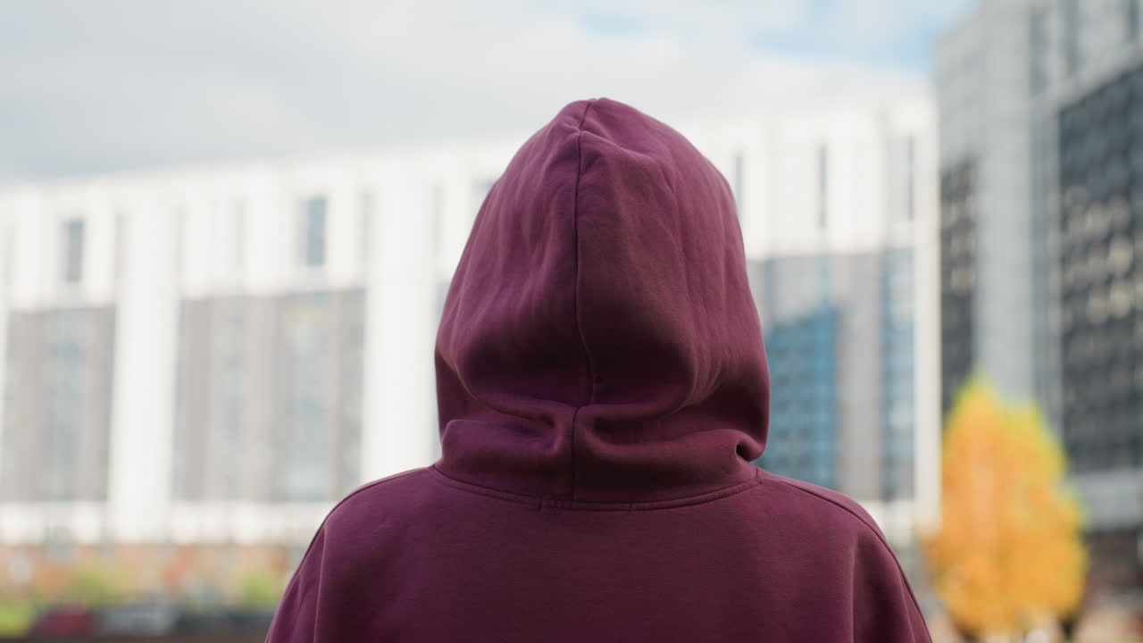 Back view of lady in wine hoodie with covered hair making fluid motion on outdoor urban pavement near modern facade subtle blur depth and silhouette and texture effect enhancing dynamic mood