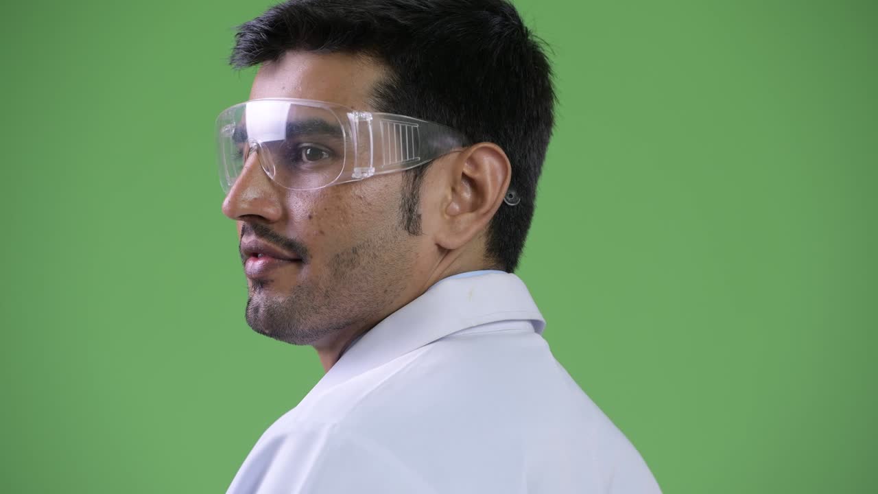 Profile view of young handsome Persian man doctor wearing protective glasses looking back