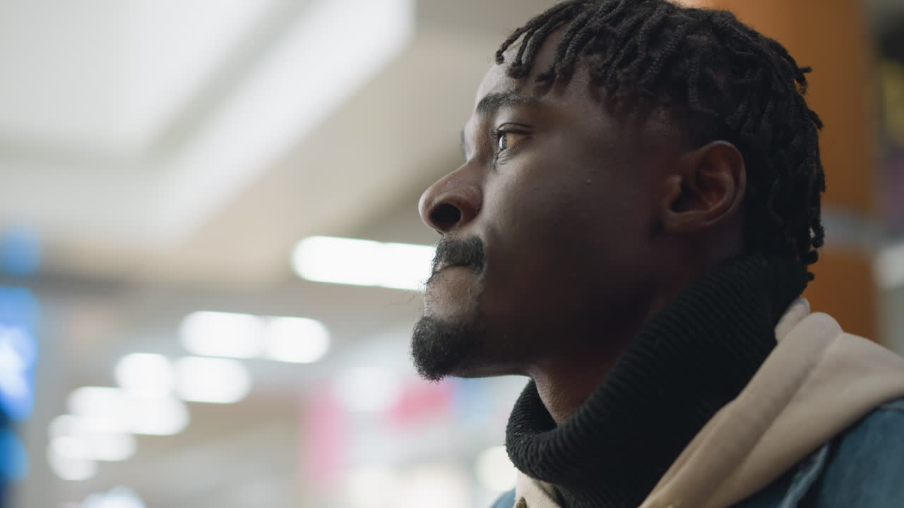 Confident young man in casual denim jacket and hoodie sipping beverage from paper cup, gazing thoughtfully ahead with focused expression, enjoying quiet moment in modern indoor setting