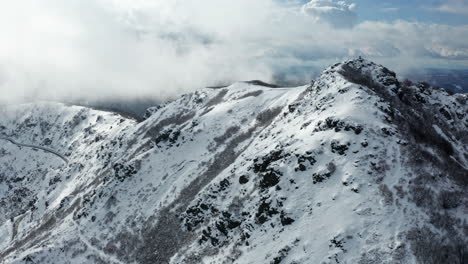 Snow-capped-mountain-peaks-with-clouds-swirling-around,-filmed-during-the-day,-aerial-view