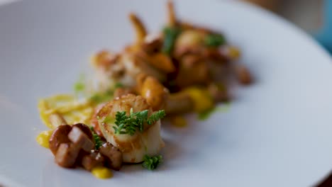 a fork lifts a seared scallop garnished with herbs from an elegant seafood dish, plated with mushrooms and sauce on a white plate in soft, natural light
