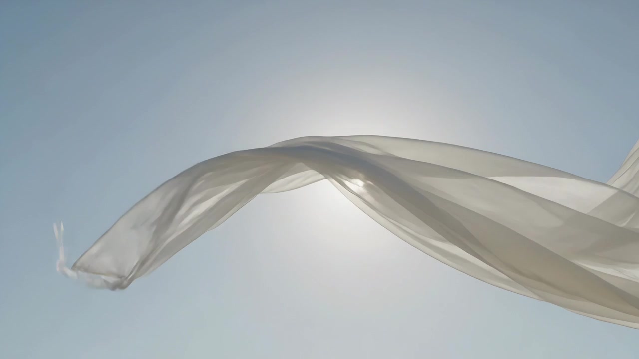 Ein schwebender, hauchdünner weißer Schal, vom Wind emporgehoben, entfaltet sich und kreuzt die helle Sonne am blauen Himmel.