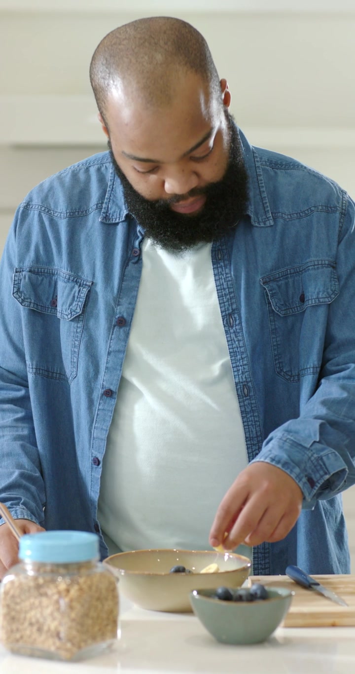 Vertical video: Adult man in denim starting by looking into bowl, picking blueberries for breakfast