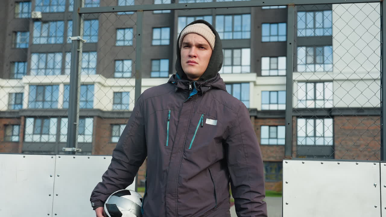 Young man in hoodie and beanie holding ball in right hand walks through bar fence with serious expression, tall office building in background