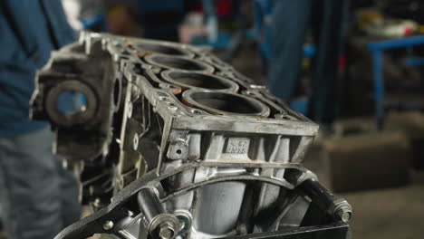 close-up of car engine with metallic details, bolts, and cylinders, placed on a stand, people in blue uniforms visible in background working in an automotive workshop filled with mechanical tools