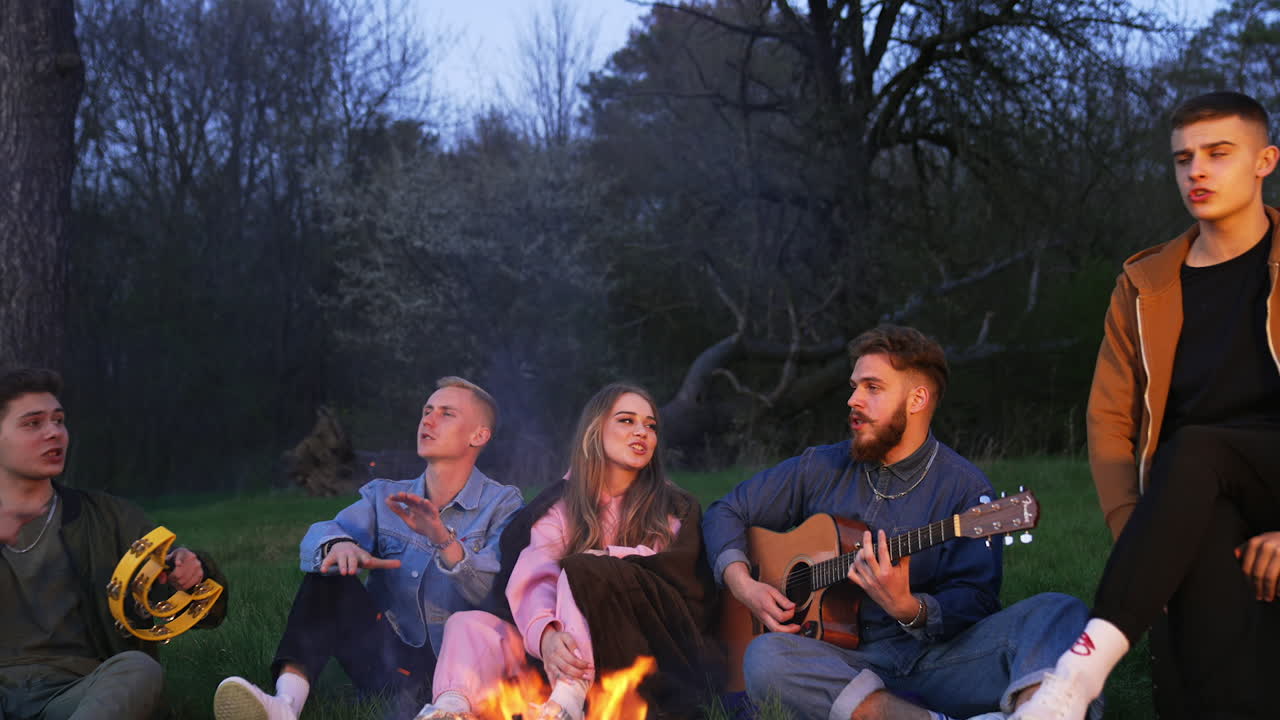 Active youth having picnic in the nature. Nice time-spending with friends, playing guitar, tambourine and singing songs together.