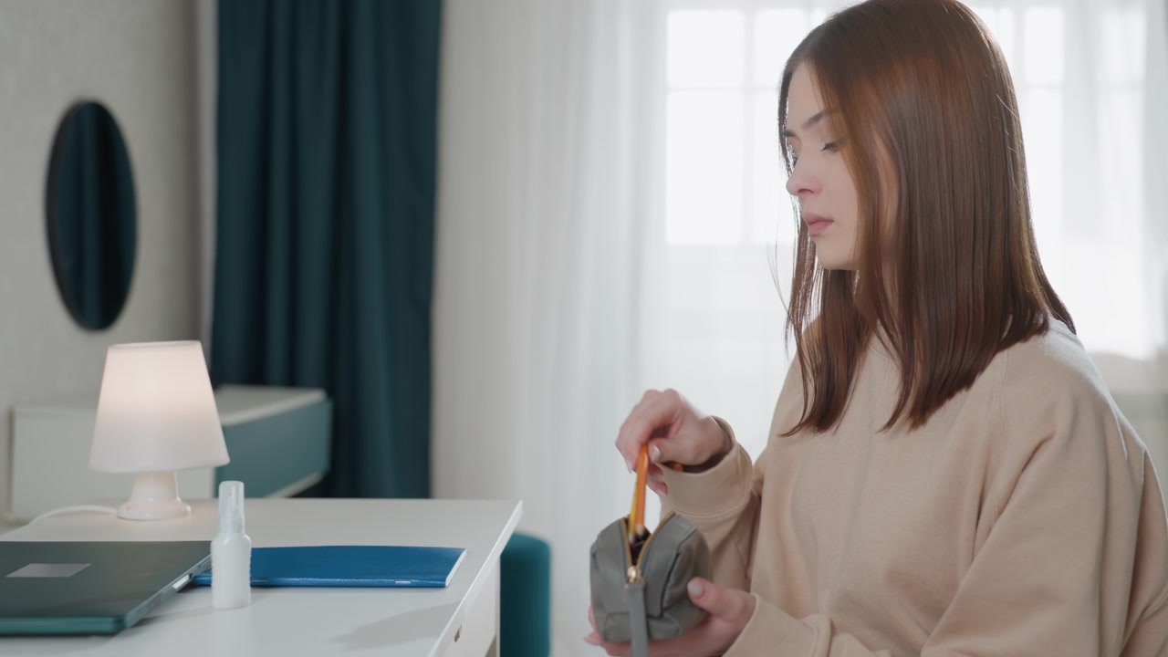 Tutor donna caucasica che conta le monete alla scrivania, scena di studio domestico tranquilla, la tutor ispeziona il salvadanaio e le monete, usa quaderno, laptop, lampada e disinfettante sulla scrivania, umore pensieroso, interno minimalista e organizzato
