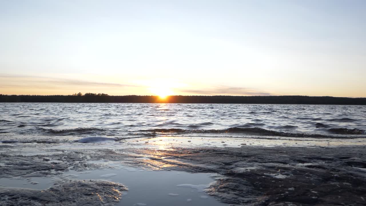 Carrellata in avanti dal basso di una spiaggia al tramonto con onde che si infrangono lentamente sulla riva, mentre la ripresa si eleva rivelando un maestoso sole che tramonta all'orizzonte