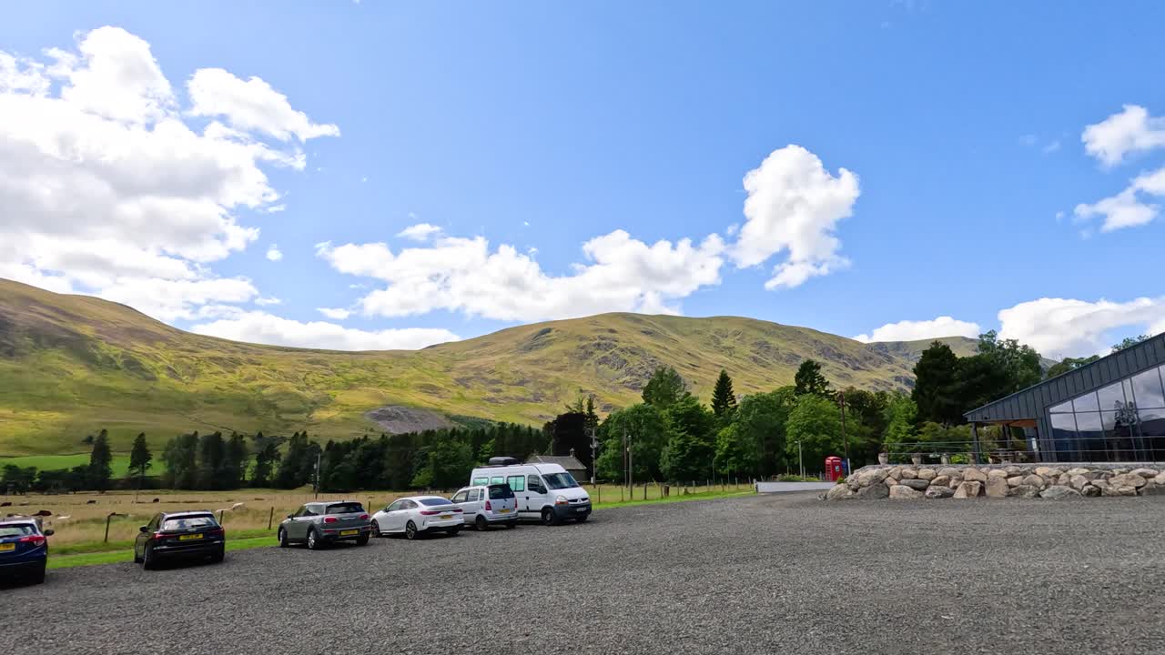 Wide-angle camera pans across a rural parking area with cars and vans, revealing rolling grassy hills, farmland, and dramatic clouds under bright daylight