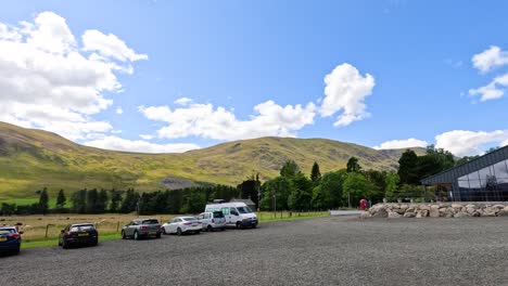 wide-angle camera pans across a rural parking area with cars and vans, revealing rolling grassy hills, farmland, and dramatic clouds under bright daylight