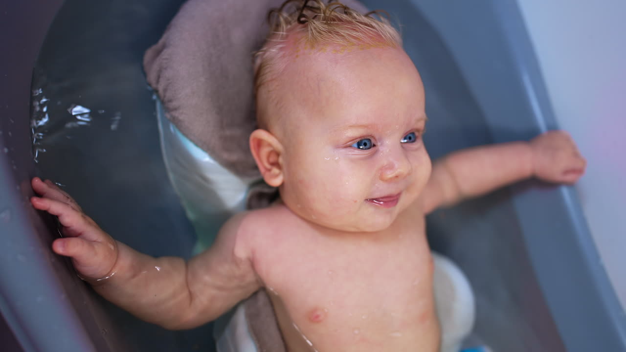 Adorable blue-eyed blond baby boy is bathed in a bath tub. Sweet kid smiles and waves his tiny hands. Close up.