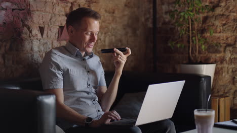 young guy talking on mobile phone and taking notes using voice assistant writing in notebook. smiling man sitting at desk with laptop