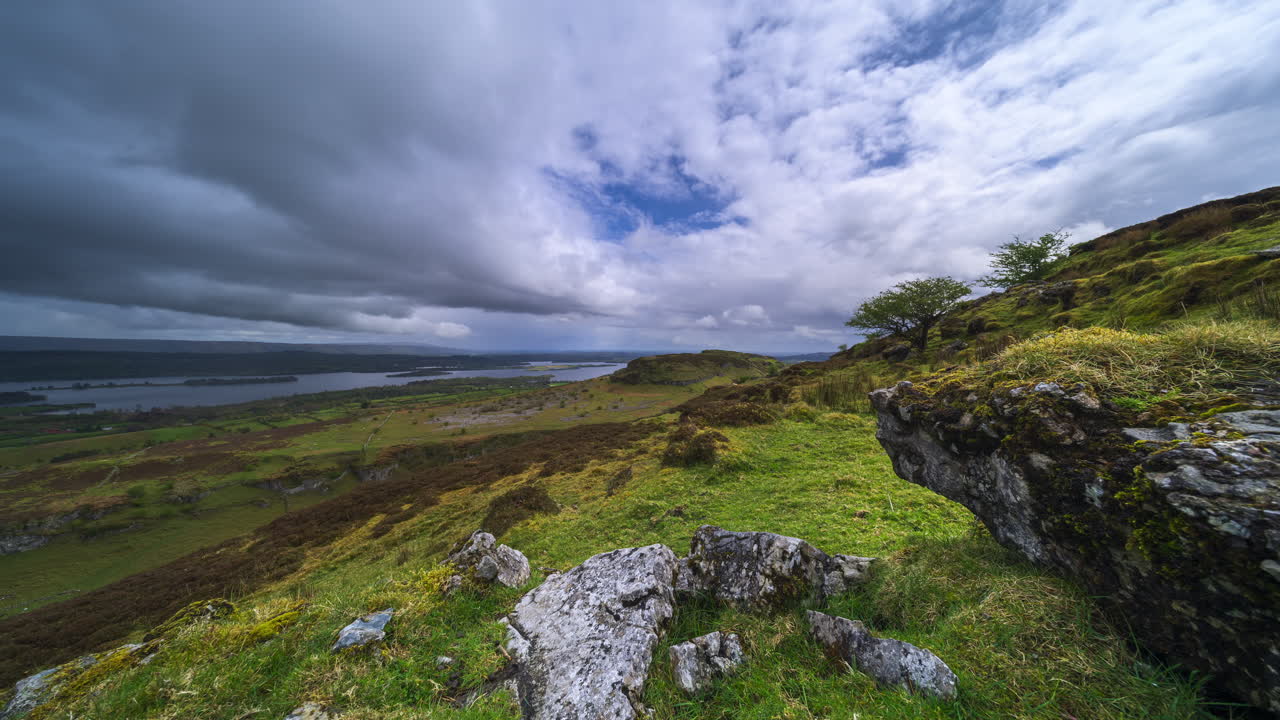 Timelapse av et landlig landskapsdal med fjellformasjon i forgrunnen og passerende regnvær, åser og innsjø i det fjerne på en vårdag sett fra Carrowkeel i County Sligo i Irland