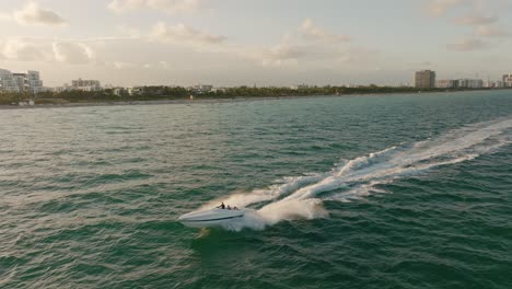 Aerial-trucking-of-people-in-a-motor-boat-sailing-in-turquoise-sea,-Miami-Beach-shore-and-resorts-in-background-at-golden-hour,-Florida,-USA