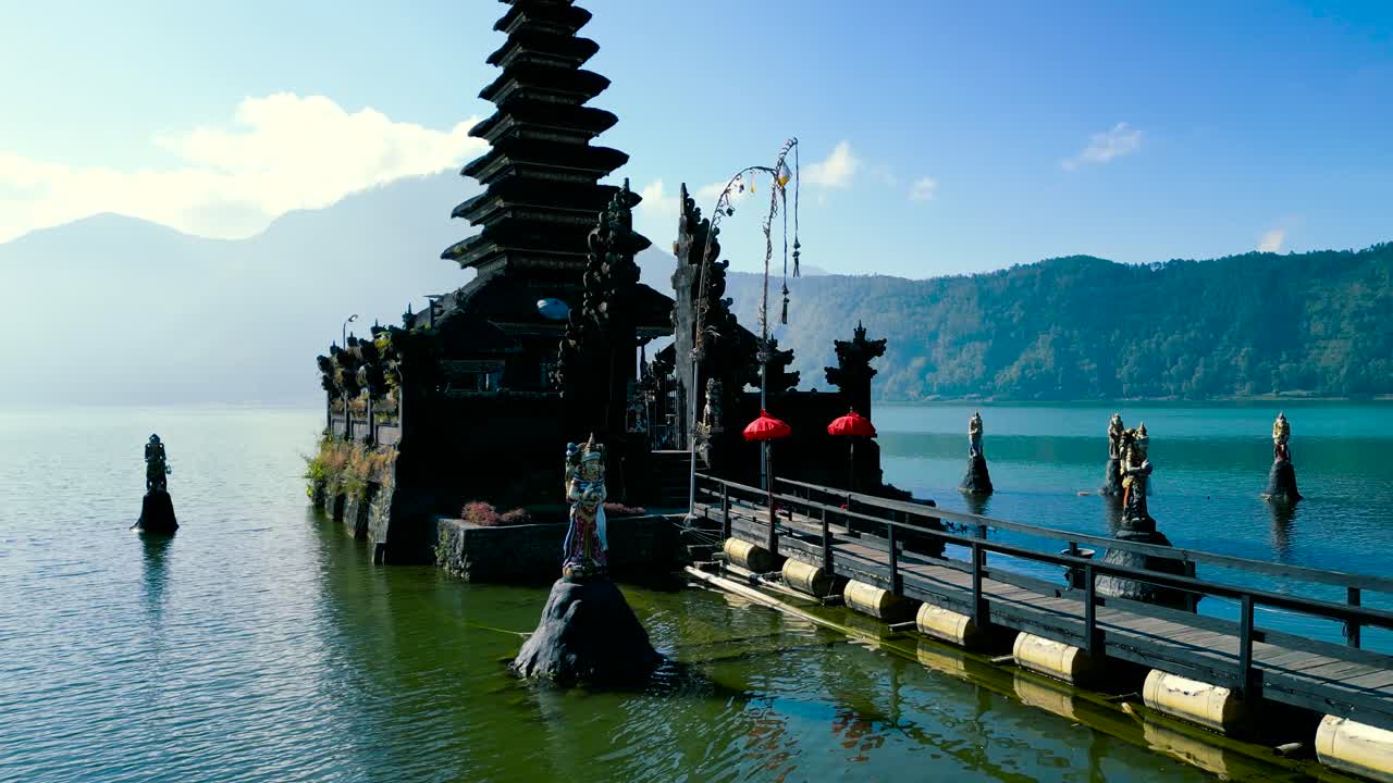 Stunning aerial footage captures Ulun Danu Beratan Temple with vibrant ceremonial umbrellas on serene Lake Beratan, Bali. Bright skies and lush mountains frame this iconic spiritual site