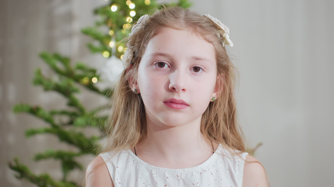 Portrait view of young girl in white gown standing in front of decorated christmas tree holding delicate white ornament in hand with thoughtful expression creating cozy festive holiday atmosphere