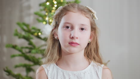 portrait view of young girl in white gown standing in front of decorated christmas tree holding delicate white ornament in hand with thoughtful expression creating cozy festive holiday atmosphere