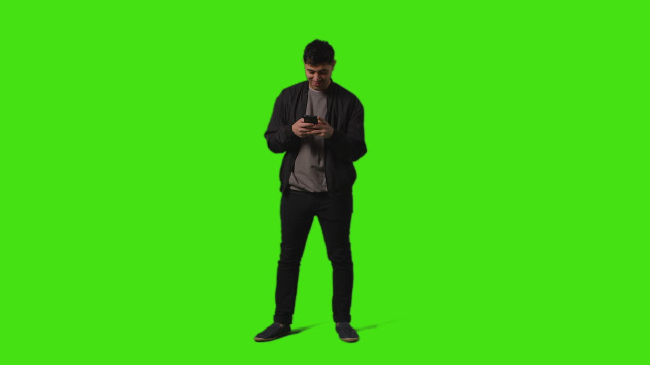 Young Man Sending Or Reading Text Message On Mobile Phone Standing Against Green Screen