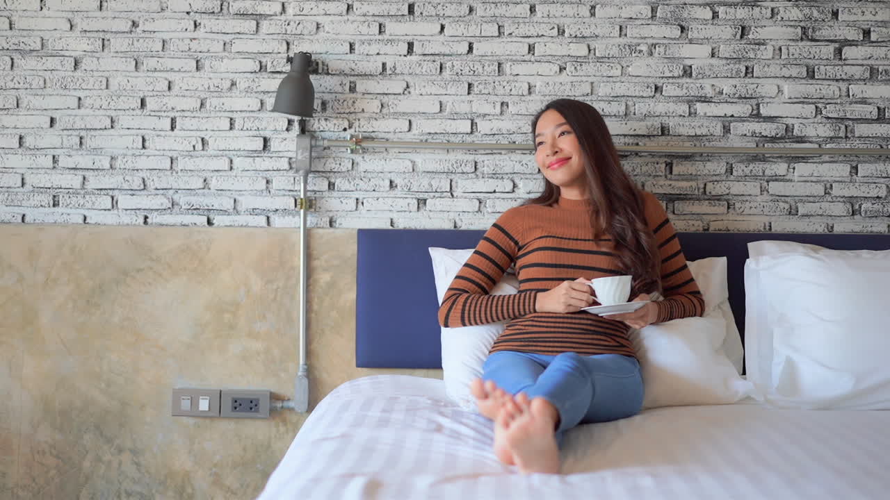 Relaxed Asian woman drinking morning coffee while lying on the bed smiling