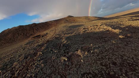 un hombre camina en una ladera volcánica rocosa bajo un arco iris vibrante en luz dorada