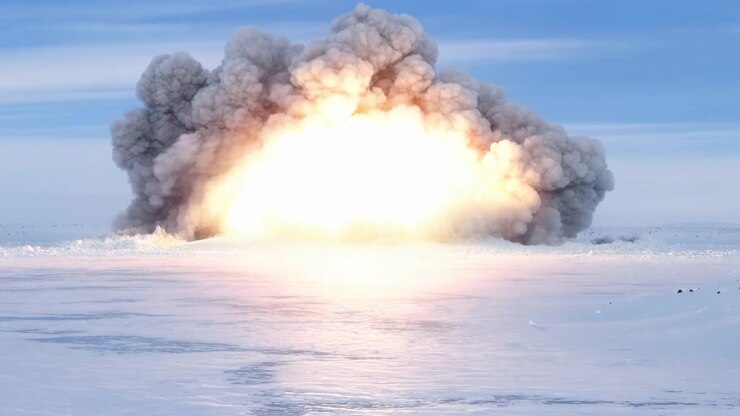 Intense orange-white flames grow at center while layered smoke spreads outward and rises above flat snowy terrain, casting warm reflections across frozen surface under clear daylight.