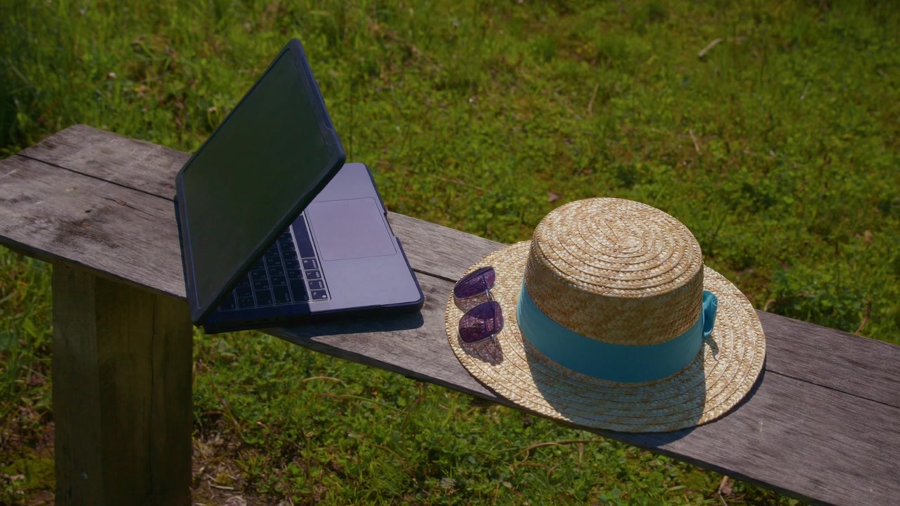 Laptop, cappello di paglia e occhiali da sole poggiano su una panca di legno. Erba soleggiata tutt'intorno. Atmosfera di lavoro all'aperto calma e rilassata. Perfetto per lavoro a distanza, viaggi o temi estivi. Ideale per blog, annunci.