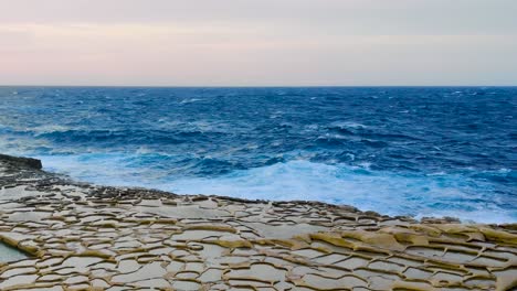 Sea-waves-crashing-into-the-coastline-with-the-salt-pans-of-Xwejni-in-Gozo,-Malta