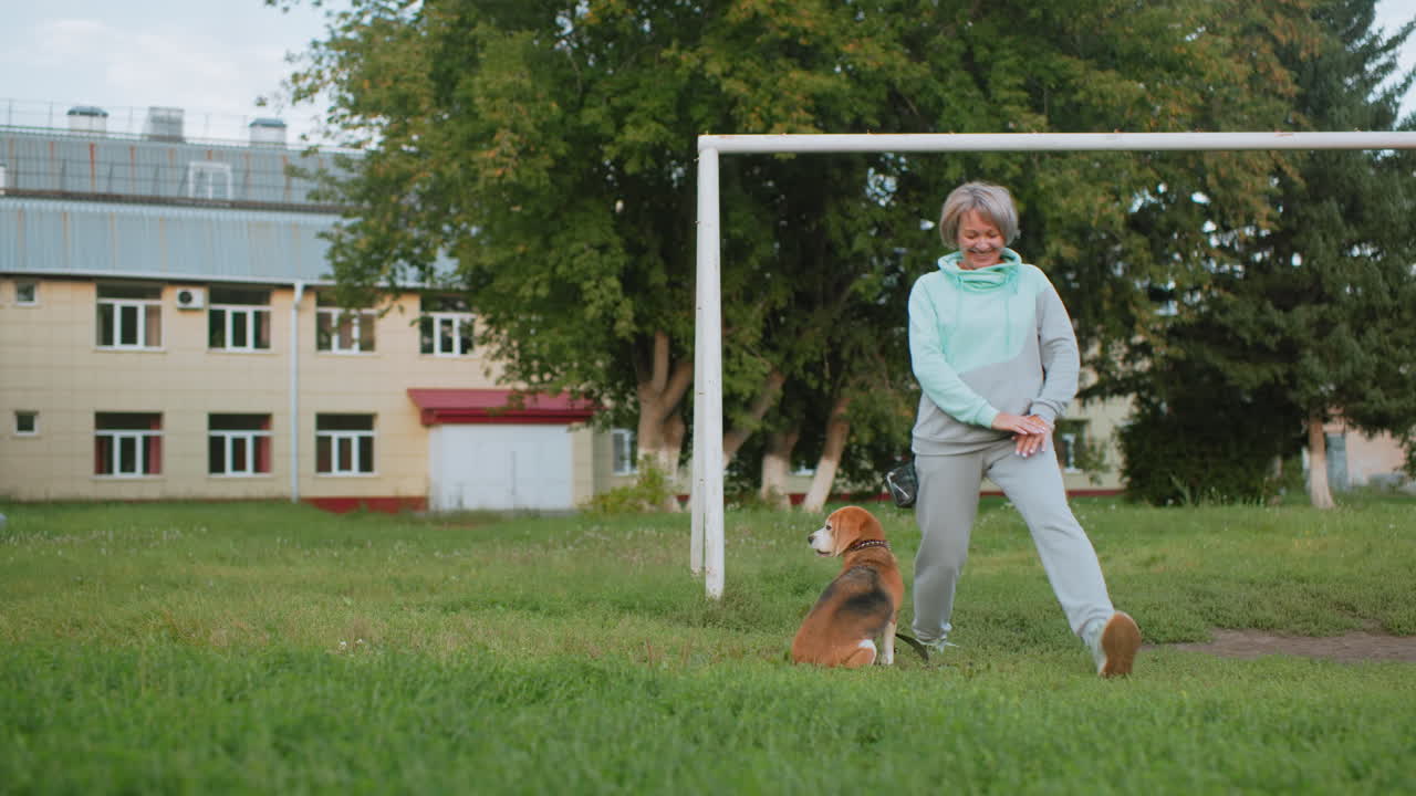 Dog tamer smiling while performing knee bend exercise on grassy field with excited bulldog jumping around playfully near goal post during outdoor session surrounded school building background