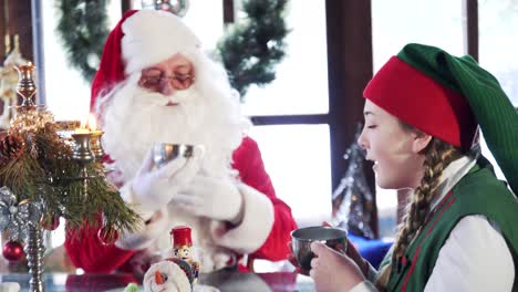 santa claus and female elf drinking tea with bagels. young woman elf with santa eat sweets at table on xmas background