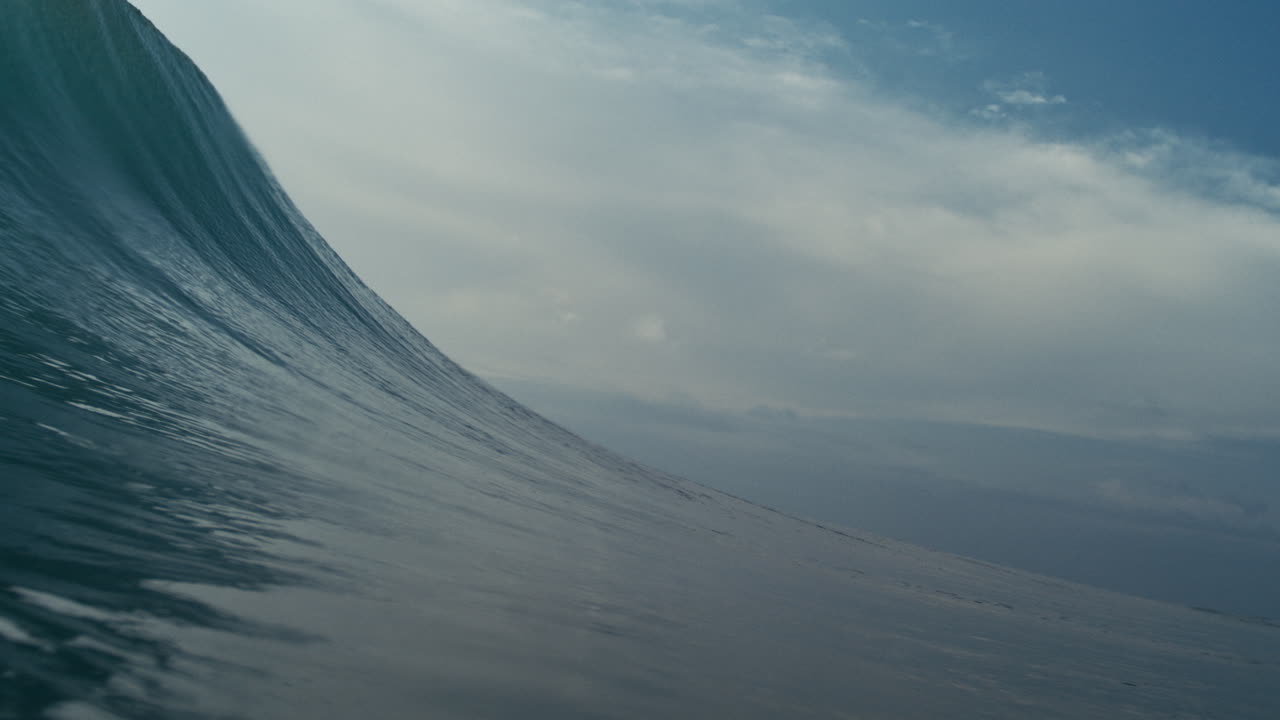 Glass like wave builds and curls under soft sky in clean open sea, shallow depth of field closeup