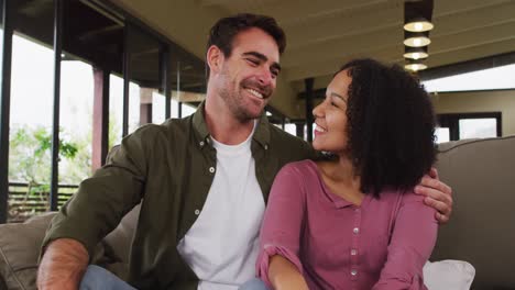 Mixed-race-couple-smiling-while-looking-at-each-other-sitting-on-the-couch-at-vacation-home