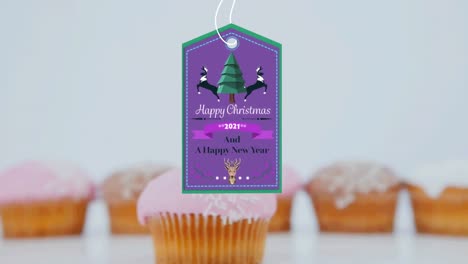 Animation-of-christmas-greetings-on-tag-over-cupcakes-on-white-background