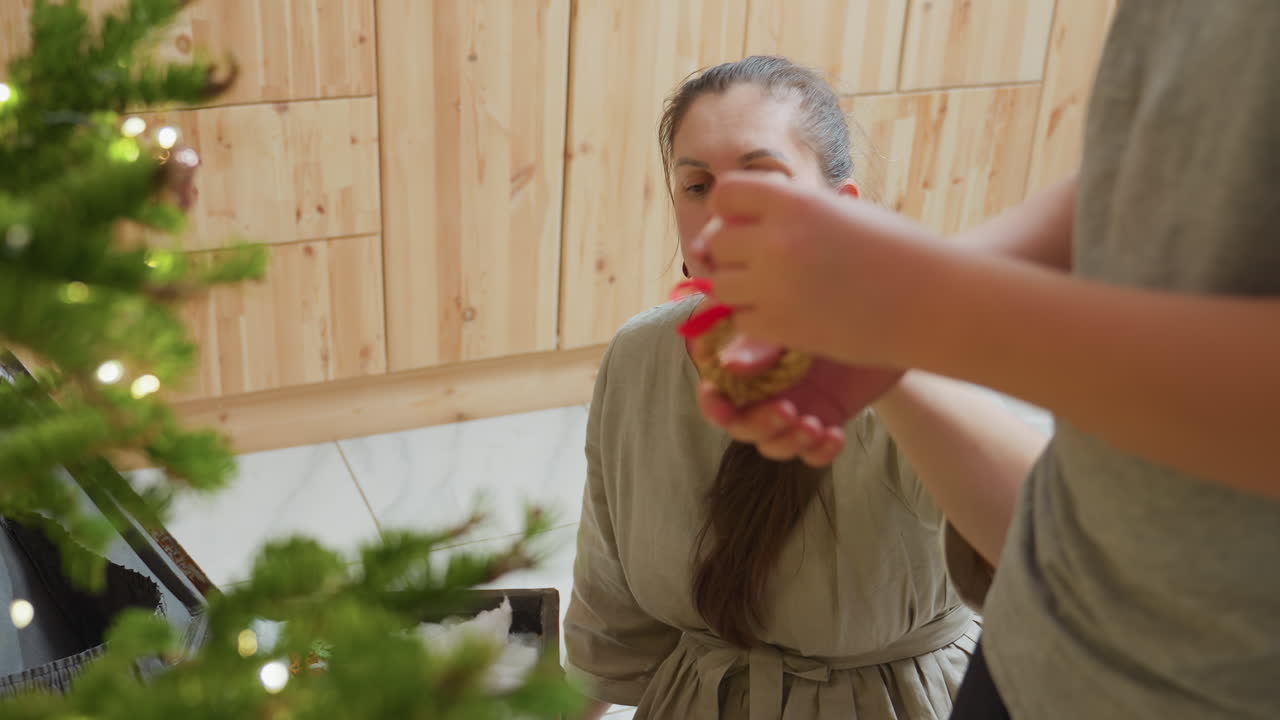 Woman picking ornament from box filled with festive decorations and handing it to boy near christmas tree, preparing for holiday celebration in cozy indoor setting with warm joyful atmosphere