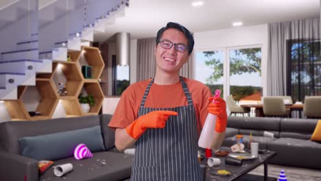 sonriente ama de casa masculina asiática con un delantal apuntando a un spray mientras está de pie en la casa