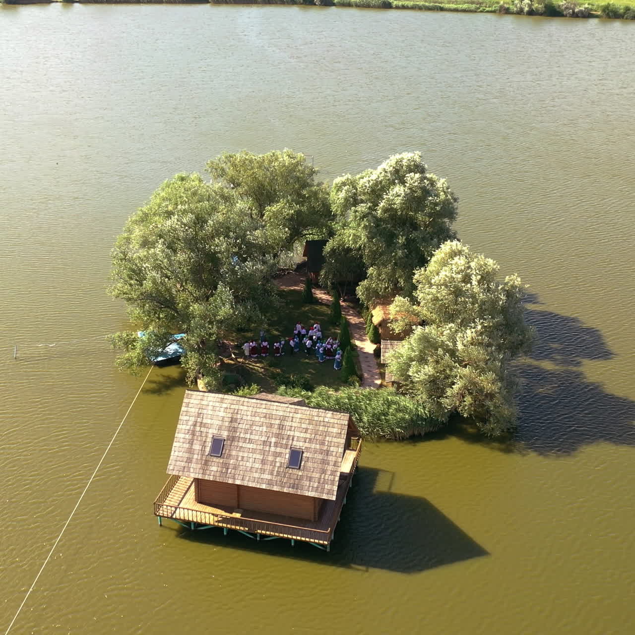 Small island with green trees and a house in the river. Aerial view of island and wooden house in the middle of a lake surrounded by water at daytime in summer. Camera moves down.