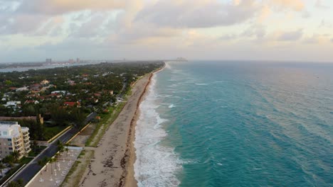 Disparo-Aéreo-De-Un-Dron-Sobrevolando-La-Isla-De-Palm-Beach-Mientras-Las-Olas-Del-Océano-Atlántico-Chocan-Contra-La-Playa-Y-El-Amanecer-Ilumina-El-Cielo