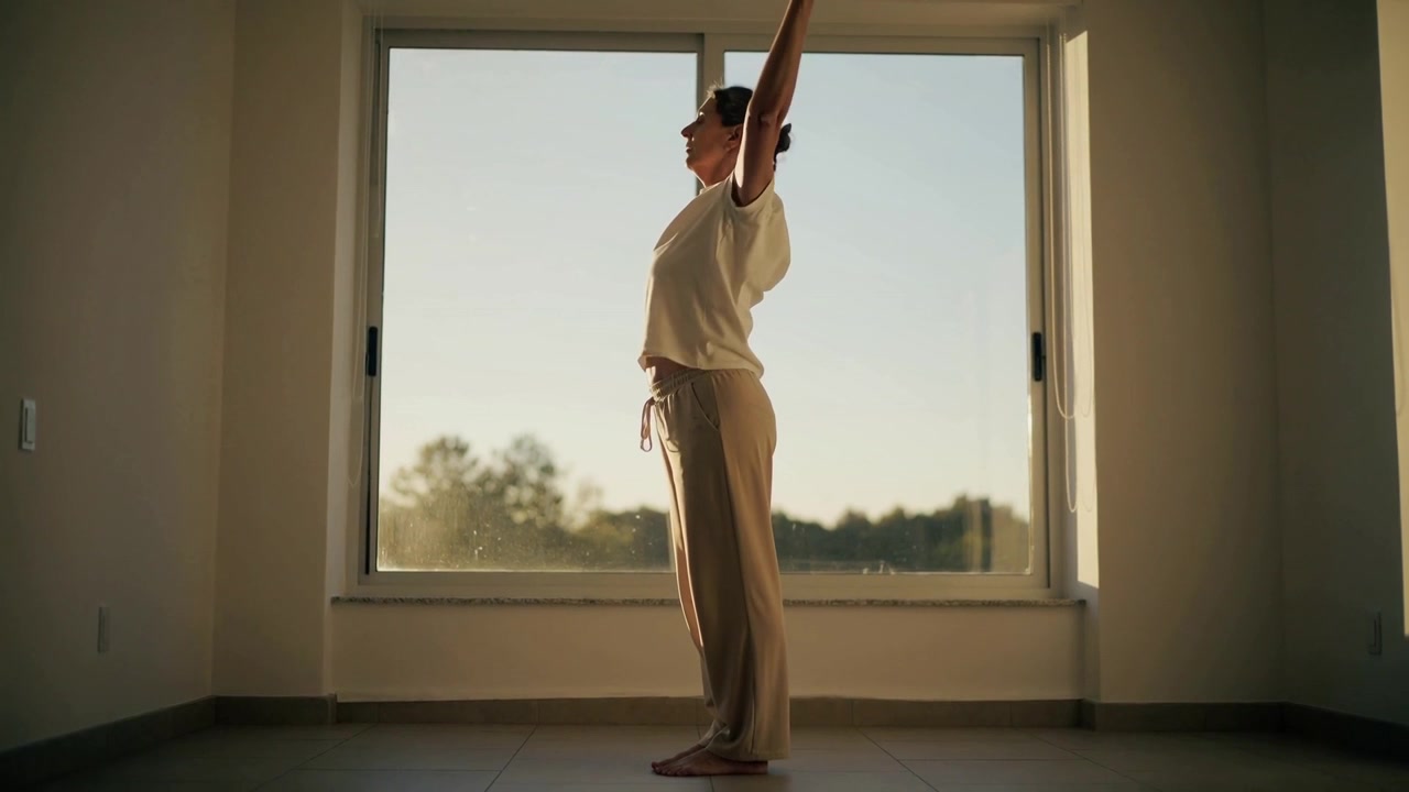 Mulher se alongando na janela, vestindo roupa de ficar em casa, estimulada pelo sol da manhã, levantando os braços, arqueando e dobrando o corpo.